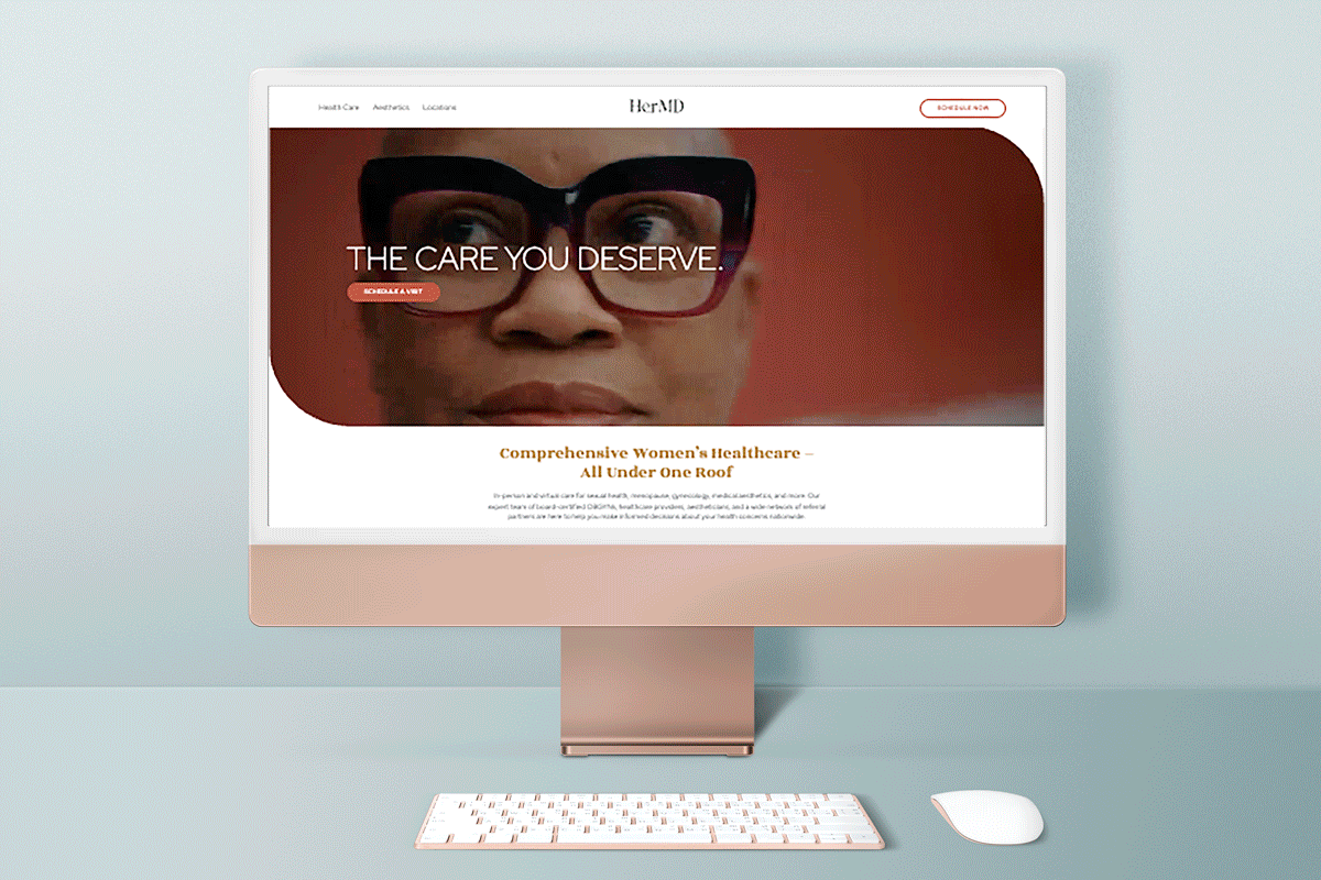 A computer screen displays the HerMD website, featuring a close-up of a womans face with glasses and the text THE CARE YOU DESERVE. The site mentions comprehensive womens healthcare. A keyboard and mouse are in front of the monitor.