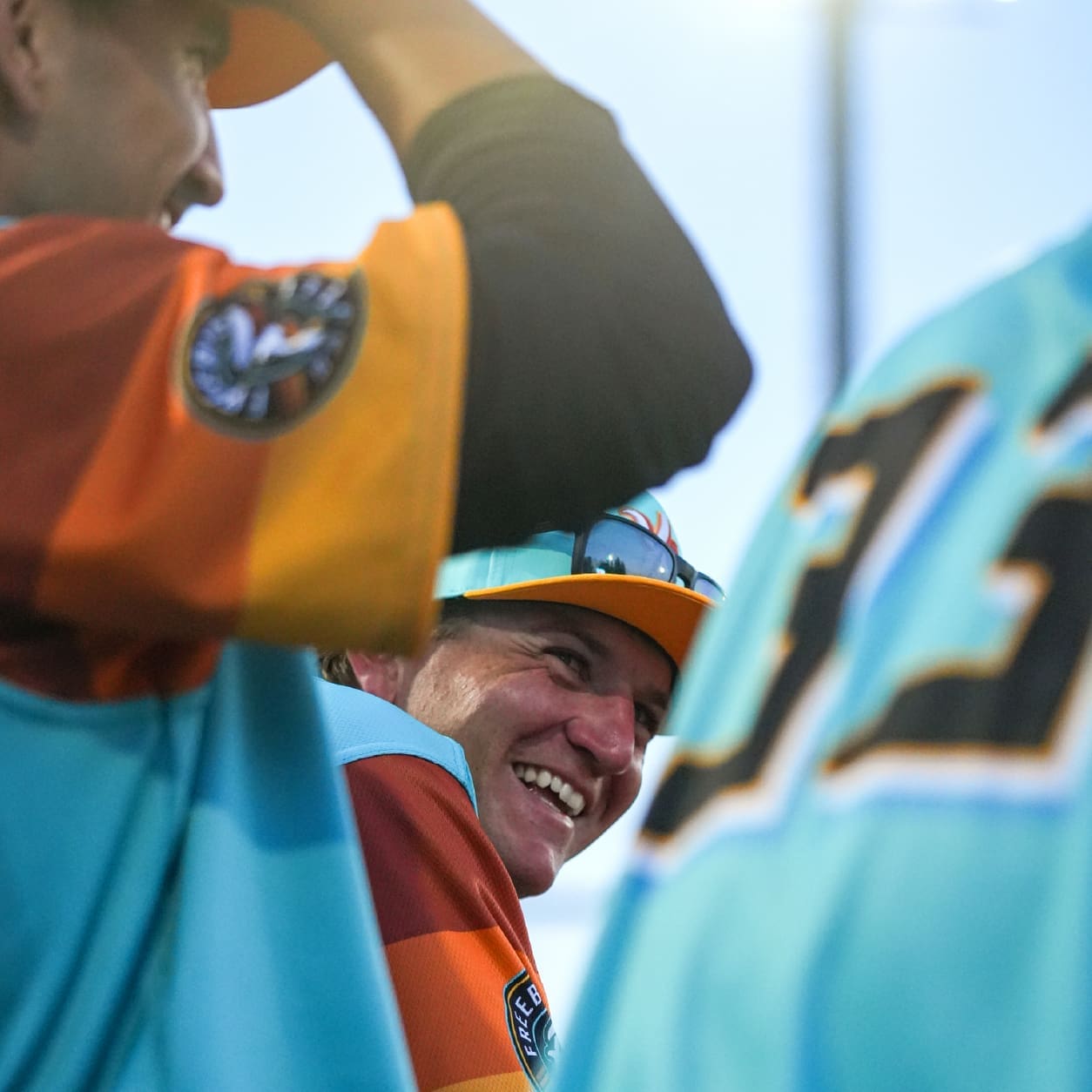 A smiling baseball player in a colorful orange, teal, and yellow uniform is seen between teammates, with sunlight shining in the background during a game.
