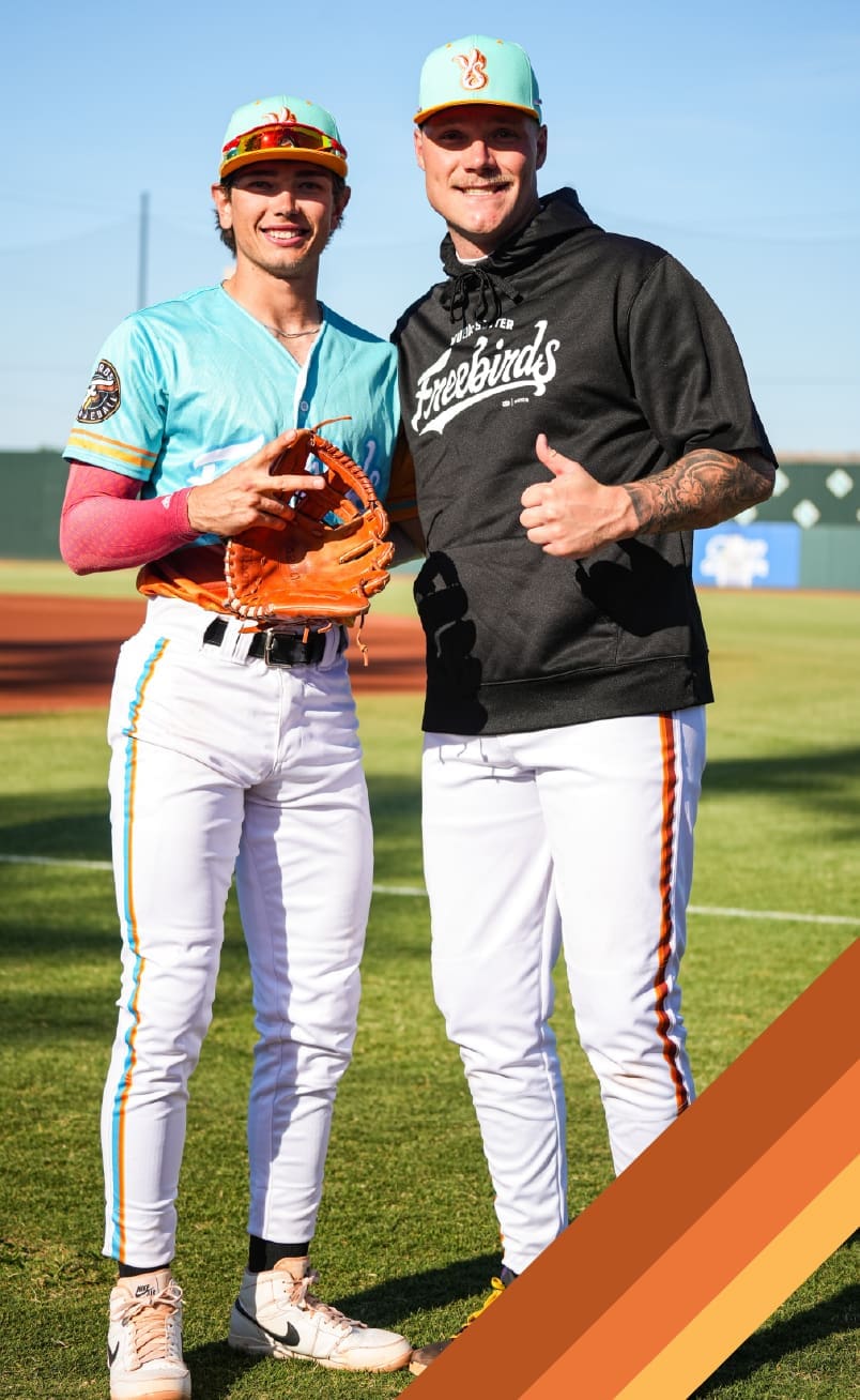 Two baseball players in colorful uniforms stand on a sunny field, smiling at the camera. One holds an orange glove and makes a peace sign, while the other gives a thumbs up. Both wear hats and white pants with orange stripes.