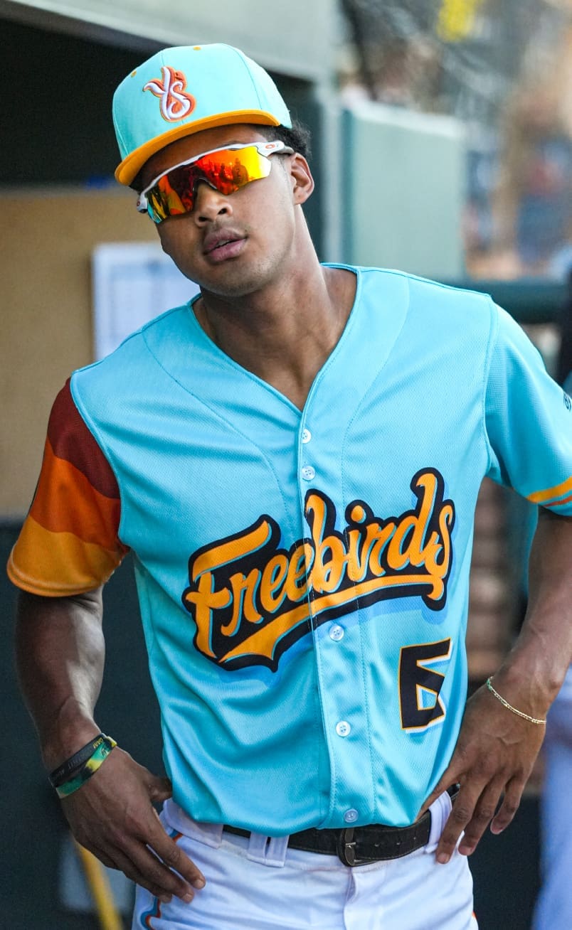 A baseball player wearing a turquoise Freebirds jersey and cap with reflective sunglasses stands with hands on hips, near a dugout.